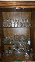 Wide shot of wooden cabinet shelves containing various crystal and glassware items including glasses, vases, plates, and bowl.