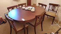 Photo showing oval wooden dining table with six chairs around it, cup and saucers on table.