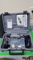 Gray Dremel rotary tool kit open showing rotary tool, wire brush attachment, guide attachment, small accessory bits in plastic box, power cord, and instruction manual inside molded carrying case.