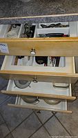 Open drawers showing assorted flatware including forks, spoons, knives and kitchen utensils such as can opener and peeler.