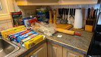 Wide view of kitchen counter showing rolling pin, black utensils, paper towel roll, wooden knife block with knives, pepper grinders, and assorted boxes of food wrap and plastic bags.
