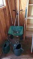 Full view of the garden tools lot inside a wooden shed, including two watering cans in foreground and spreader, rake, and wooden handled tools in background.