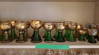 Full shelf view showing 11 German wine goblets with green ribbed stems and gold decorated bowls.