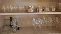 Shelf showing clear coffee cups with handles on saucers, and several clear glass wine glasses with stems in front. Also visible crystal serving plates and bowls with intricate patterns.