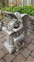 Front side view of the silver painted kneeling angel lawn ornament showing chipping on wings and hair, placed on a brick patio near flower bed.
