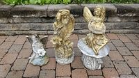 Three weathered lawn statues on a brick patio in front of a raised garden bed: one small Grecian-style, two angels with silver finish