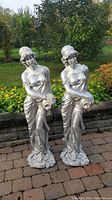 Two tall Grecian women lawn statues in silver, standing outdoors on brick pavers in front of garden and bushes.