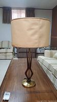 Front view of lamp showing beige fabric shade with braided trim and sculptural teak and brass base.