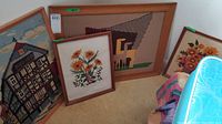 Four framed needlepoint and yarn artwork pieces arranged on the floor showing various themes including a building, floral designs, and geometric pattern with horses.