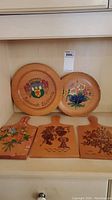 Five hand painted wooden items displayed: three rectangular bread boards with floral and bird motifs, two round plates with floral bouquet and coat of arms designs.