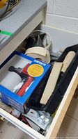 Open drawer photo showing assorted painting tools including Krebs 35T sprayer box, paint rollers, paint tray, masking tape, and other accessories.