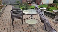 Full view of patio set on brick patio including a round glass tabletop, umbrella folded with cover, six stacked metal chairs, and a smaller round side table.
