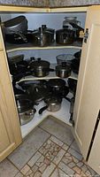 Photo showing multiple pots, pans, and lids arranged inside corner cabinet with beige doors and tiled floor.