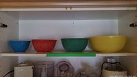 Four nested Pyrex mixing bowls in blue, red, green, and yellow on a white shelf.