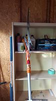 Full length view of the extendable orange pole tree lopper inside a shed leaning against shelving showing the curved cutting blade at the top and rope control system.