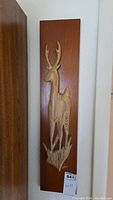 Front view of the wood carving mounted on a wooden plaque showing the deer shape with antlers and surrounding grass detail.
