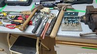 Photo shows two wooden handled hammers, adjustable wrench, pliers, some snips, and other metal hand tools spread on a tabletop.