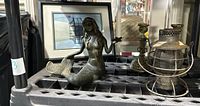 Metal mermaid statue holding something in her hand, two brass candle holders are positioned behind her and a framed lighthouse painting is in the background.