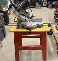 Front view of the Delta compound miter saw mounted on a wooden table with red metal legs, showing the miter gauge and blade guard.