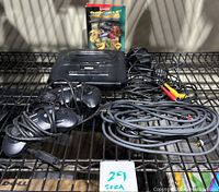 Photo showing black Sega Genesis console with two wired controllers, connection cables, and Street Fighter 2 Special Champion Edition game cartridge on metal shelf with identification tag.
