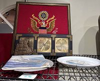 Framed fabric wall hanging of The Great Seal, Springfield 3702 barometer/thermometer/humidity meter set with wooden frame, metal ship bookends, and two decorative plates on display shelf