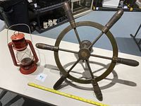 Full image of the vintage brass ship wheel next to red Dietz junior lantern on table with measuring tape.