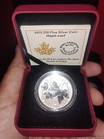 Front view of silver coin showing maple leaf design inside a protective capsule, placed in red velvet box with certificate in background