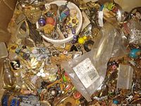 Top-down view of a cardboard box containing assorted vintage costume jewelry pieces, small figurines, beads, pins, and other small objects mixed together, highlighting variety and colors.
