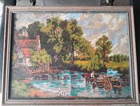 Front view of vintage framed needlepoint artwork depicting rural landscape with water, trees, and buildings.