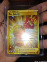 Front view of Toy Catcher gold sparkle holo Pokemon Trainer card held in hand inside protective plastic sleeve, showing full art and text clearly.
