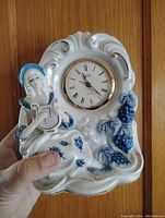 Porcelain figurine clock held by a hand in front of a wooden background, showing frontal and side views of the clock and detailing of figure and grapes.