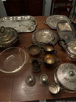 Wide shot of the lot on wood table showing various brass, copper, glass, and silver-tone serving trays, tureen, cups, candleholders