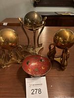 Front view of all three brass globes on distinct stands and the red hand-painted trinket dish arranged on a wooden surface.