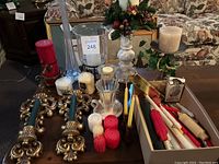 Wide view showing large tall glass vase, pair of ornate gold-tone metal wall candle holders with blue taper candles, tall cream-colored candle holder, crystal-style candle holder, and numerous red, white, and cream candles of various sizes.