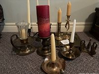 Overview of six metal candlestick holders with candles of varied sizes and styles on floor near wall.