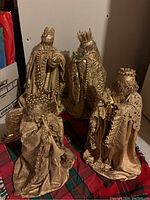 Group photo of four figurines – three wise men and one angel dressed in gold fabric and lace, standing on a red plaid cloth.