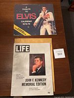 Photo shows the John F. Kennedy Memorial Edition LIFE magazine and Elvis Presley 1978-79 Tribute calendar side by side on a wooden surface with a numbered lot card.