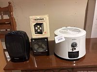 Three heaters/fans on a table: white Sunbeam heater, black Classic heater, Microfurnace heater in box.