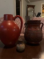 Photo showing three pottery jugs placed on a table: a large red jug, a medium brown decorative jug, and a small multicolored jug.
