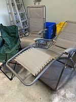 Pair of beige reclining orbital loungers with metal frames and fabric seats, shown in reclined and upright positions inside a garage with ladders and recycling bins visible.
