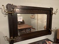 Full view of wooden framed mirror mounted on wall with six ornate brass hooks attached on sides and top edges.