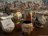 Photo showing five decorative pitchers arranged on table with floral couch background, visible details of shapes and colors