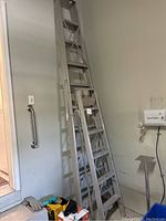 Two aluminum stepladders leaning against a garage wall next to a door and some garage items.