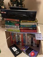 Front view of Sharp VC-A5833 VHS player on top of multiple VHS tapes and a VHS headcleaner tape, image shows remote control on player and titles of tapes visible.