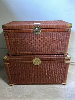 Two stacked wicker storage trunks showing woven wicker texture and brass latches on corners and front.