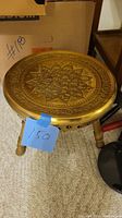 Photo of round brass foot warmer on carpet with blue price tag '150'. Shows decorative embossed top and wooden legs.
