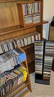 Music CDs stored in wooden racks and plastic holder on floor and shelves. Titles mostly visible on spines.