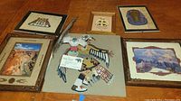 Photo showing multiple framed artworks and decorative items arranged on wood surface. Includes Native American and Egyptian themed art, photos of canyons, fabric panel with bird design, and parrot figurine.