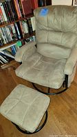 Glider chair and ottoman in taupe ultra suede fabric with signs of wear, on wood floor next to bookshelf.