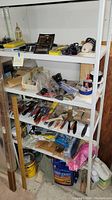 White metal shop shelf filled with assorted hand tools, power tool accessories, hardware, and shop supplies on four shelves.